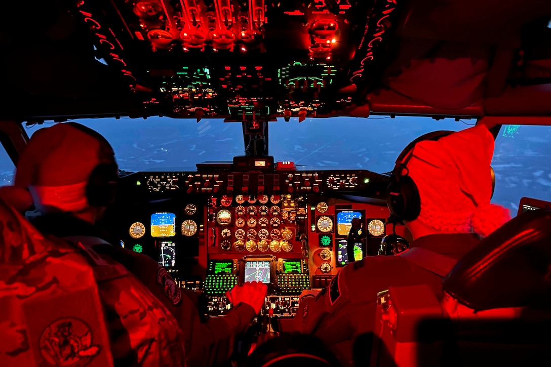 Two airmen ride in the colorful cockpit of an airborne aircraft bathed in red light.