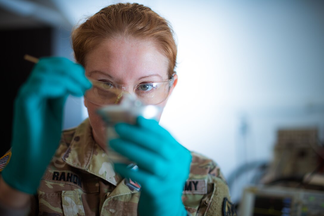 A soldier wearing safety glasses and rubber gloves holds up and inspects an object in a laboratory.