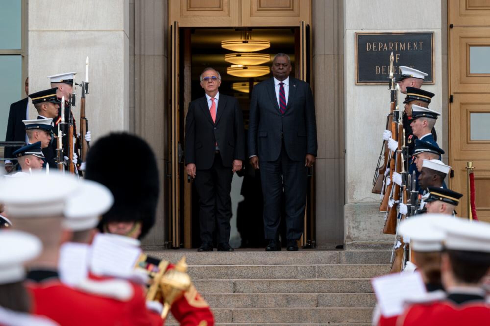 Two men stand at the top of the steps of a building overseeing an honor guard and a military band. The sign on the building indicates that the are at the Pentagon.
