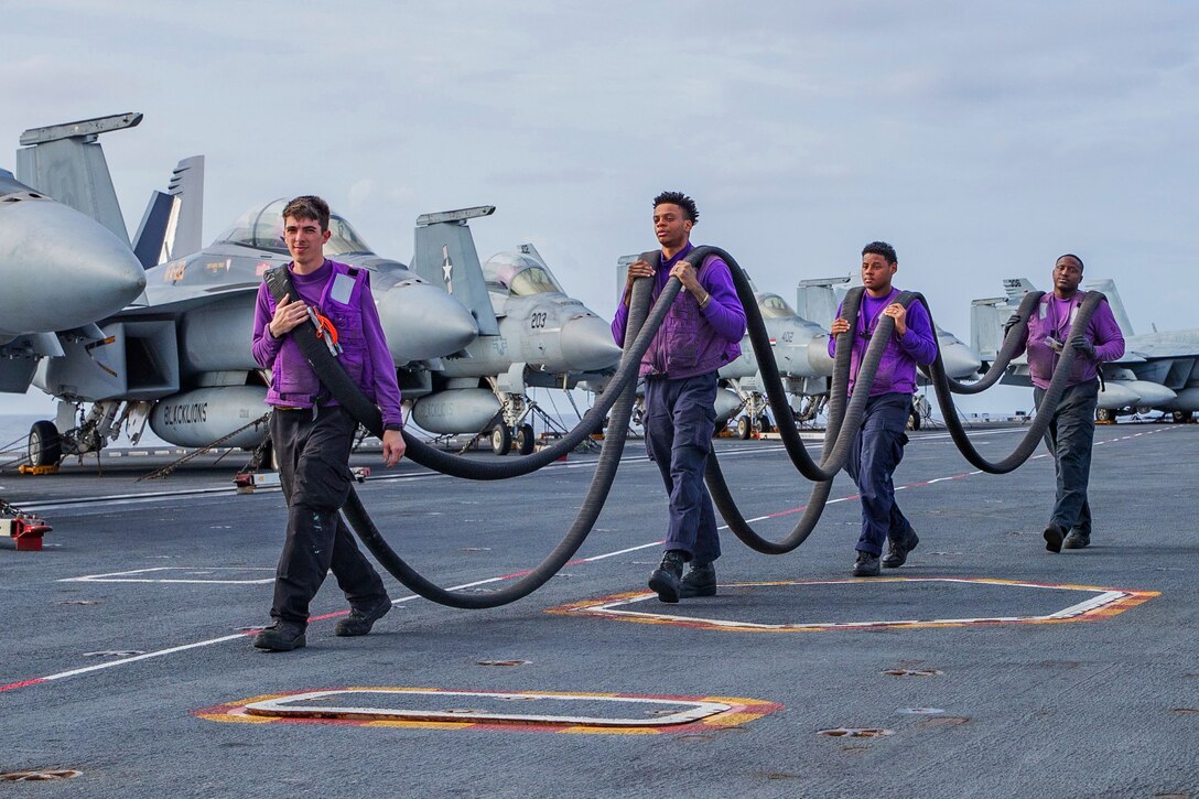 Four sailors walk in a single line and carry a fuel hose over their shoulders.