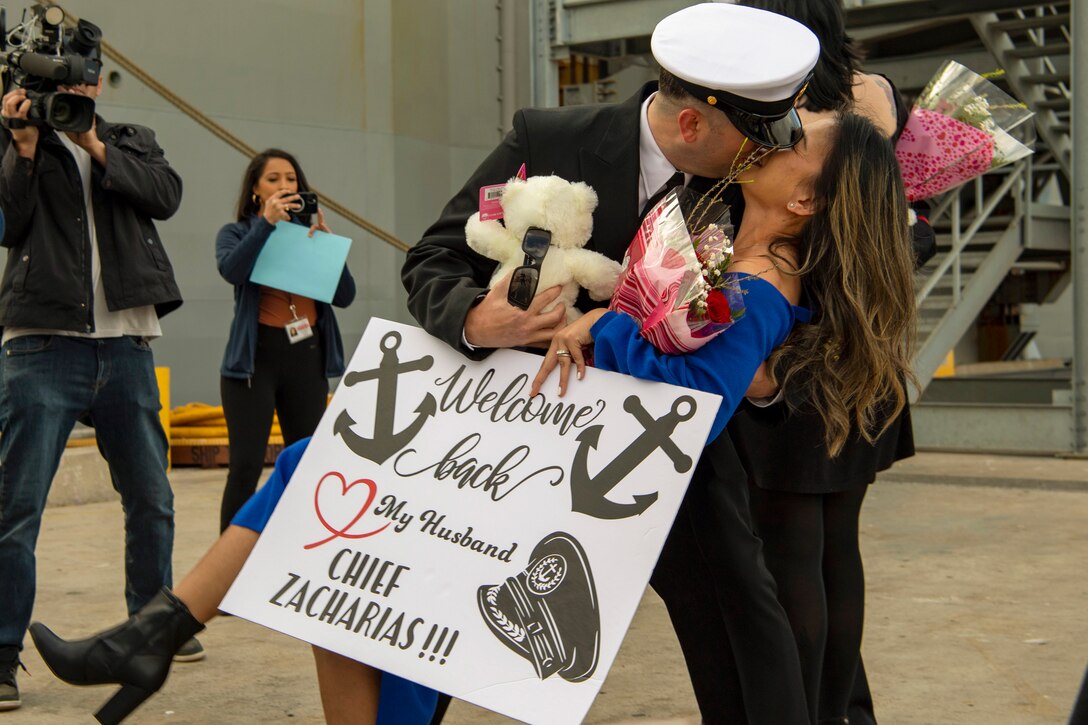 A sailor dips and kisses his wife as people record with a camera and a phone.