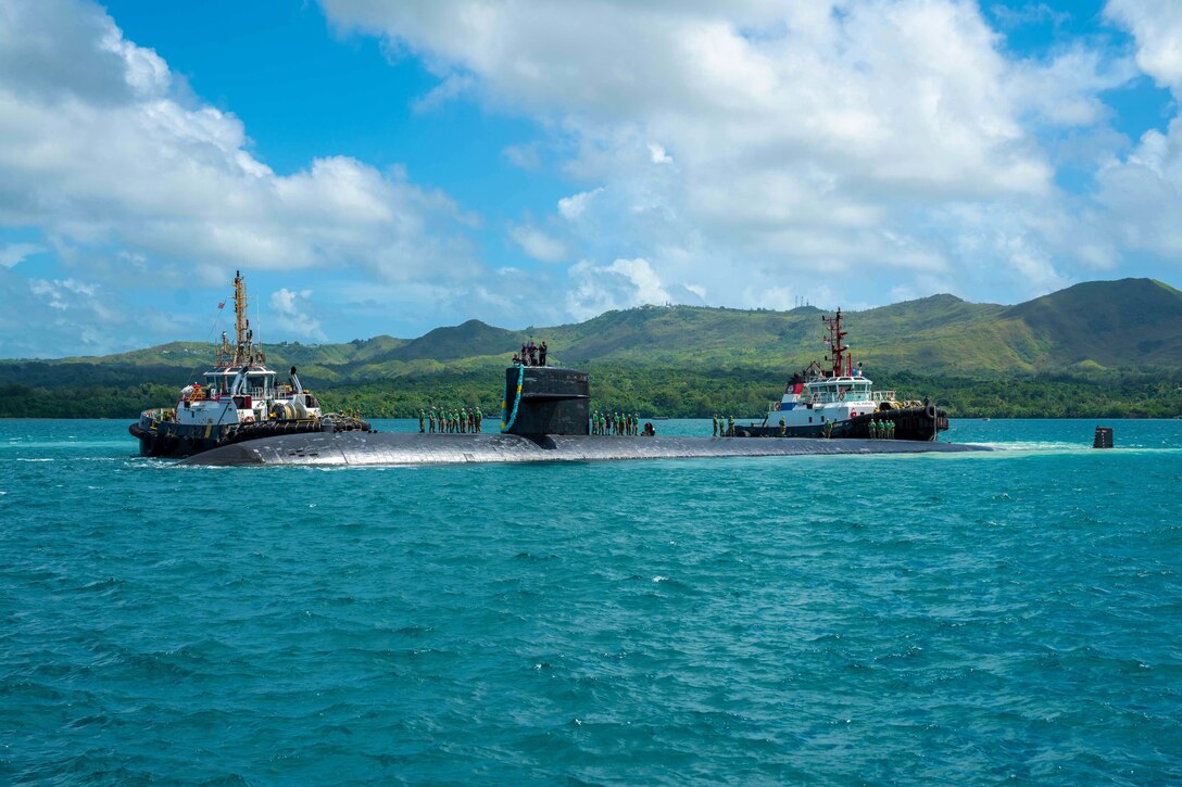 Crew members stand on a submarine that's flanked by a boat at each end.