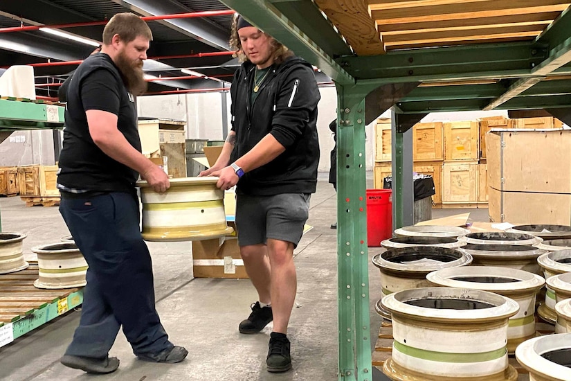 Two men carry an aircraft wheel in a warehouse.