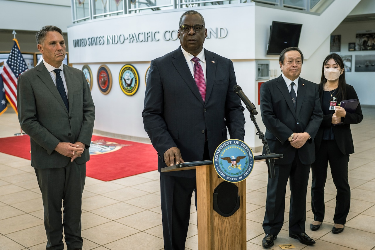 A man stands at a podium, as two men and a woman look on.