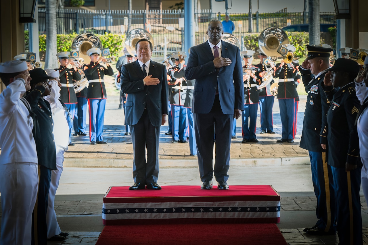 During a ceremony, two men stand side by side with their hands over their hearts. A military band is in the background.