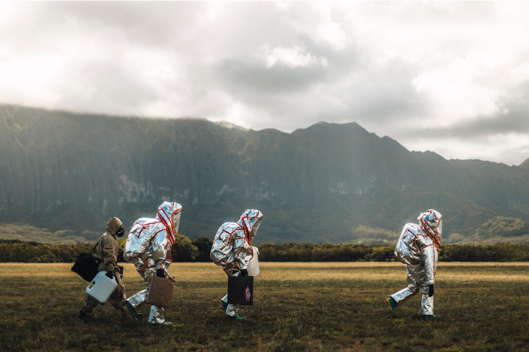 Marines wearing protective gear carrying bins walk through a field with mountains in the background.