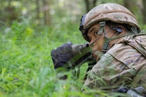 An Airman crouched in the grass peers out with binoculars