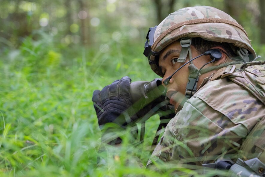 An Airman crouched in the grass peers out with binoculars