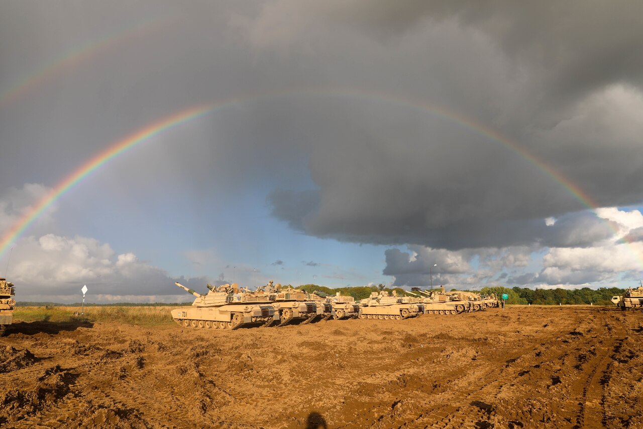Soldiers in tanks in an open field train. A rainbow is in the sky.