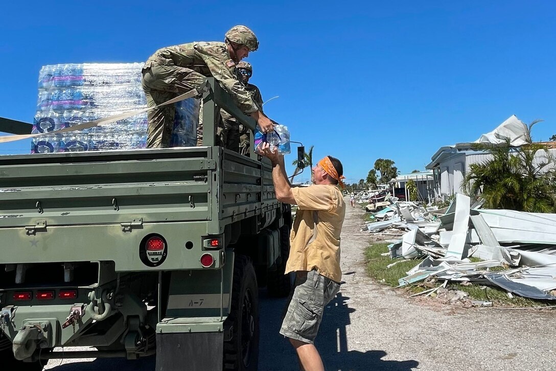 A soldier in the back of a military pickup loaded with bottled water passes a case to a person on the ground.