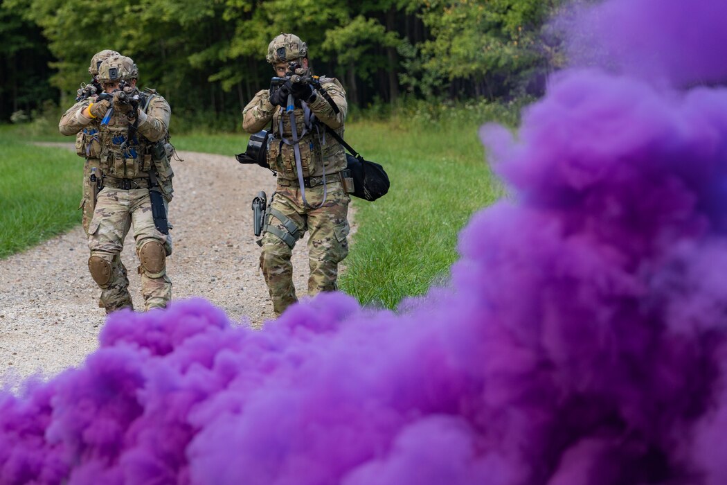Airmen in combat gear walk towards a smog of purple smoke.