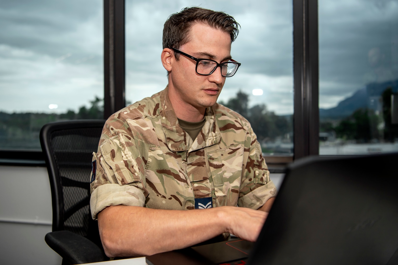 Man in United Kingdom military uniform wearing glasses, types on a laptop