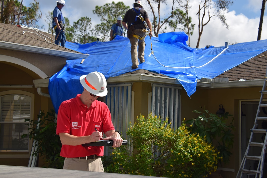 Workers apply a blue tarp to the roof of a damaged house while a man in hard hat and red shirt takes notes on the ground.