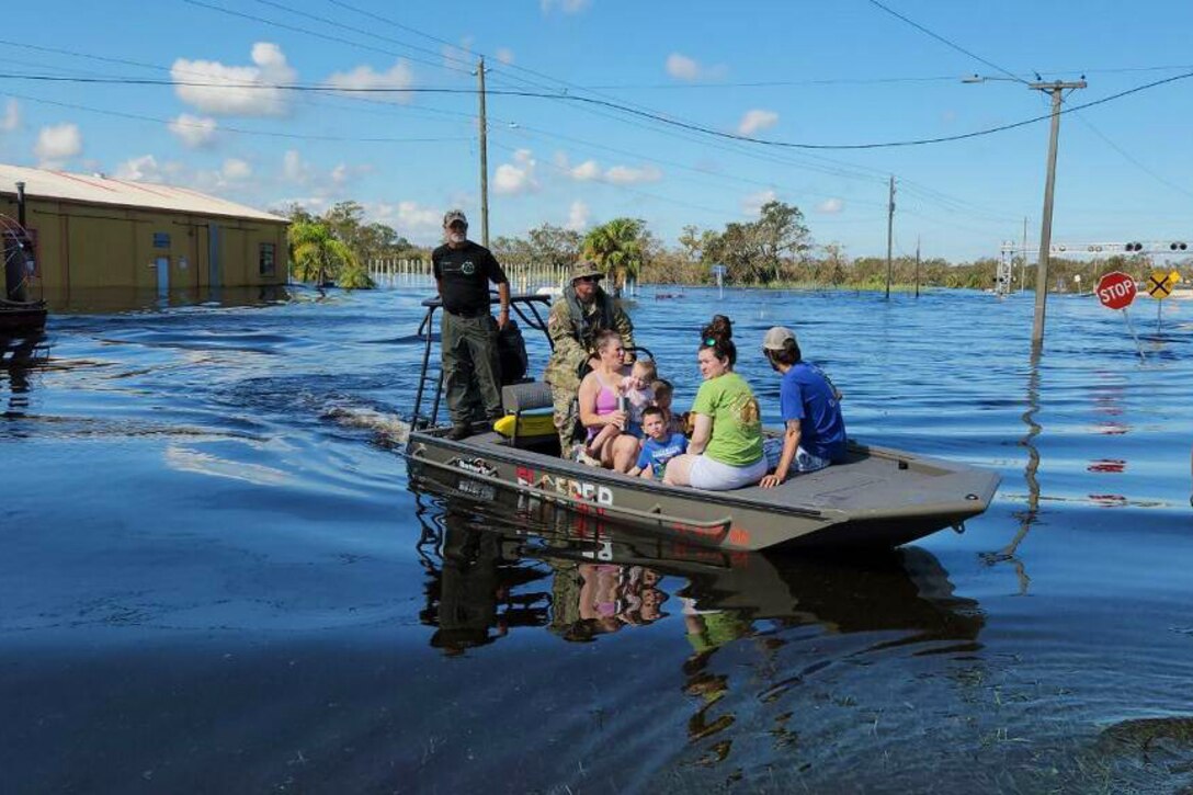 A group of residents and a service member sit on a boat traveling through flood waters.