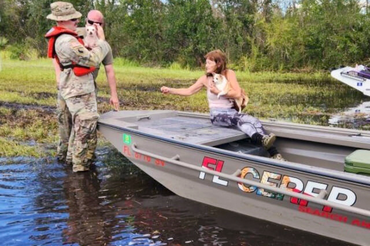 A resident sits in a small boat holding a dog as two guardsmen stand next it holding a dog.