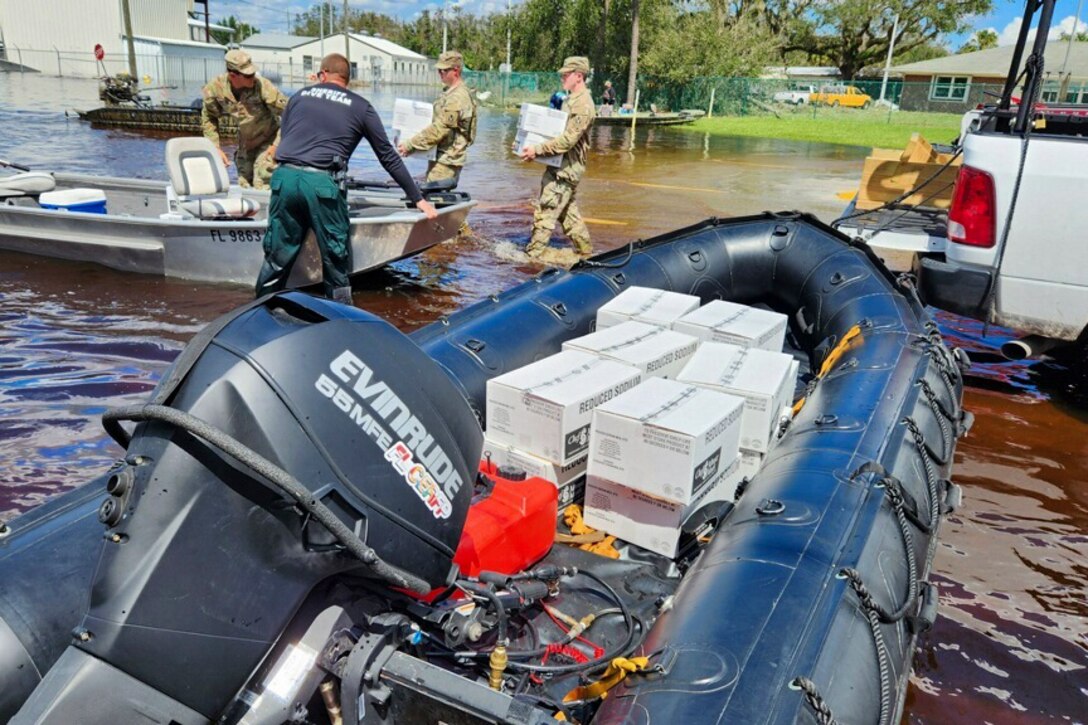 Guardsmen carry white boxes from a truck into small boats in a flooded lot.