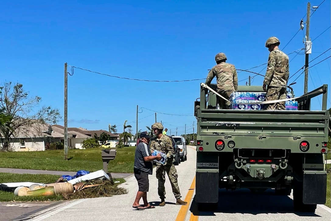 A soldier hands a person a case of water as other soldiers stand inside a military vehicle.