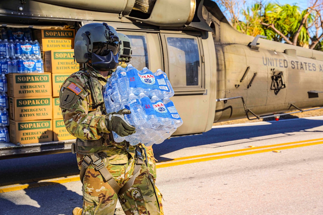 A soldier carries two cases of water from a parked helicopter.