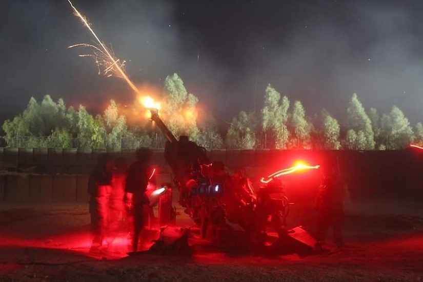 A large gun fires at night, leaving a trail of sparks.