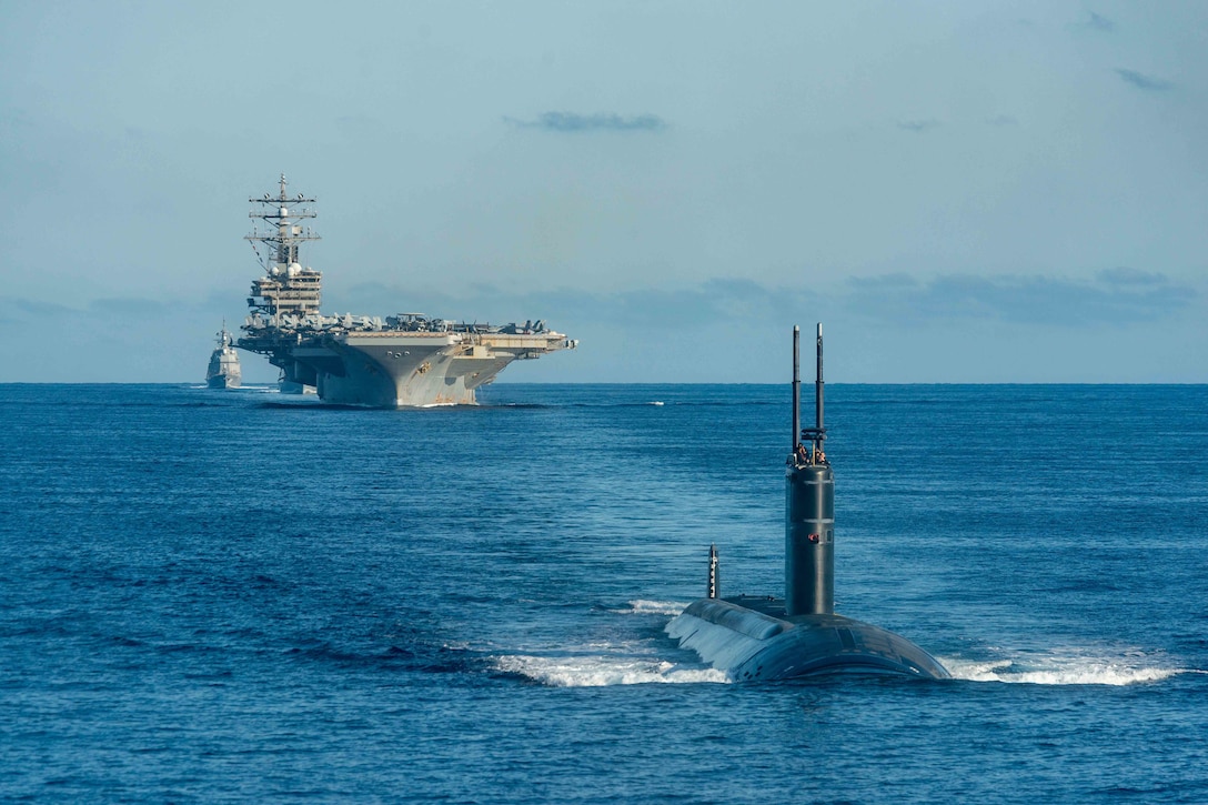 A submarine steams in front of an aircraft carrier and some other ships.