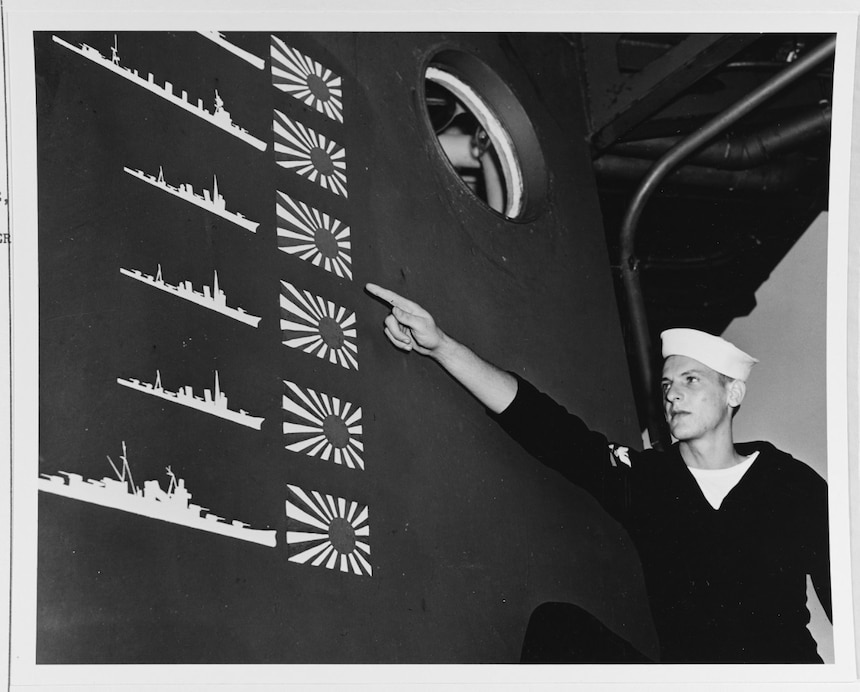Sailor W.R. Martin points out details of the Japanese trophy flags painted on the cruiser's pilothouse as a scoreboard of enemy ships claimed sunk in the Battle of Cape Esperance, 11-12 October 1942. The six Japanese ships (two heavy cruisers, a light cruiser and three destroyers) represented in this scoreboard greatly overstates the actual enemy losses, which were one heavy cruiser (Furutaka) and one destroyer (Fubuki) sunk and one heavy cruiser (Aoba) badly damaged. This overclaiming was typical of contemporary night surface actions. Photographed at the Philadelphia Navy Yard, Pennsylvania, soon after Boise arrived there for battle damage repairs in November 1942. Official U.S. Navy Photograph, now in the collections of the National Archives. (NHHC 80-G-36296)