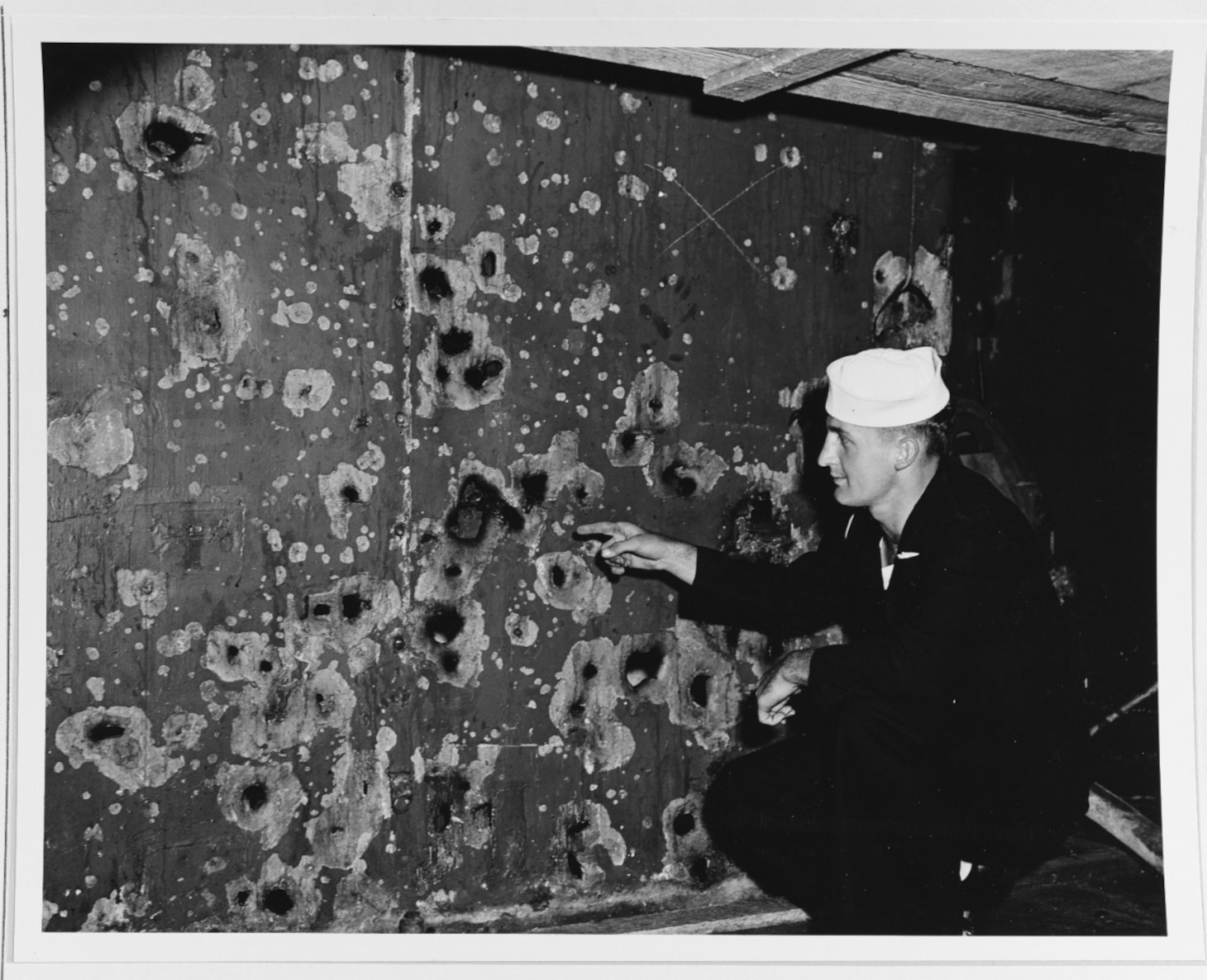 Seaman First Class Charles Olinger points out shell fragment damage to the ventilation trunk at the front of the superstructure, just above the main deck. Photographed at the Philadelphia Navy Yard in November 1942. This damage was received during the Battle of Cape Esperance, 12 October 1942. It was caused by a shell that struck the face of turret # 3 and detonated on impact. The explosion was about thirty feet away from the damage seen here. Official U.S. Navy Photograph, now in the collections of the National Archives. (NHHC 80-G-36296)