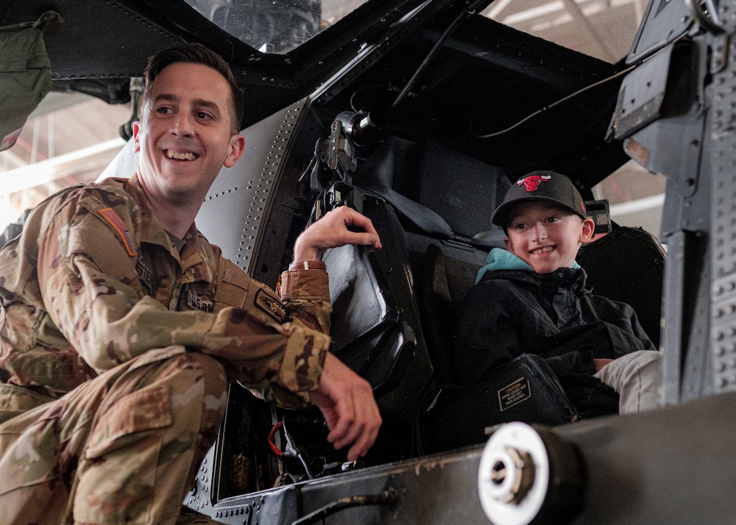 U.S. Army Capt. Bryan Westervelt (left), Apache pilot, Headquarters and Headquarters Company, 12th Combat Aviation Brigade, smiles for a photo with a young boy sitting inside an AH-64D Apache Longbow helicopter in hangar two on Katterbach Kaserne, Germany, April 21, 2022. The static display was in conjunction with a speaker series event discussing the importance of collective security and the success of the transatlantic partnership and friendship. 12 CAB is among other units assigned to V Corps, America's Forward Deployed Corps in Europe. V Corps works alongside NATO Allies and regional security partners to provide combat-ready forces, execute joint and multinational training exercises, and retain command and control for all rotational and assigned units in the European Theater. (U.S. Army photo by Staff Sgt. Thomas Mort)