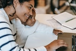 A woman holds her baby while working on a laptop