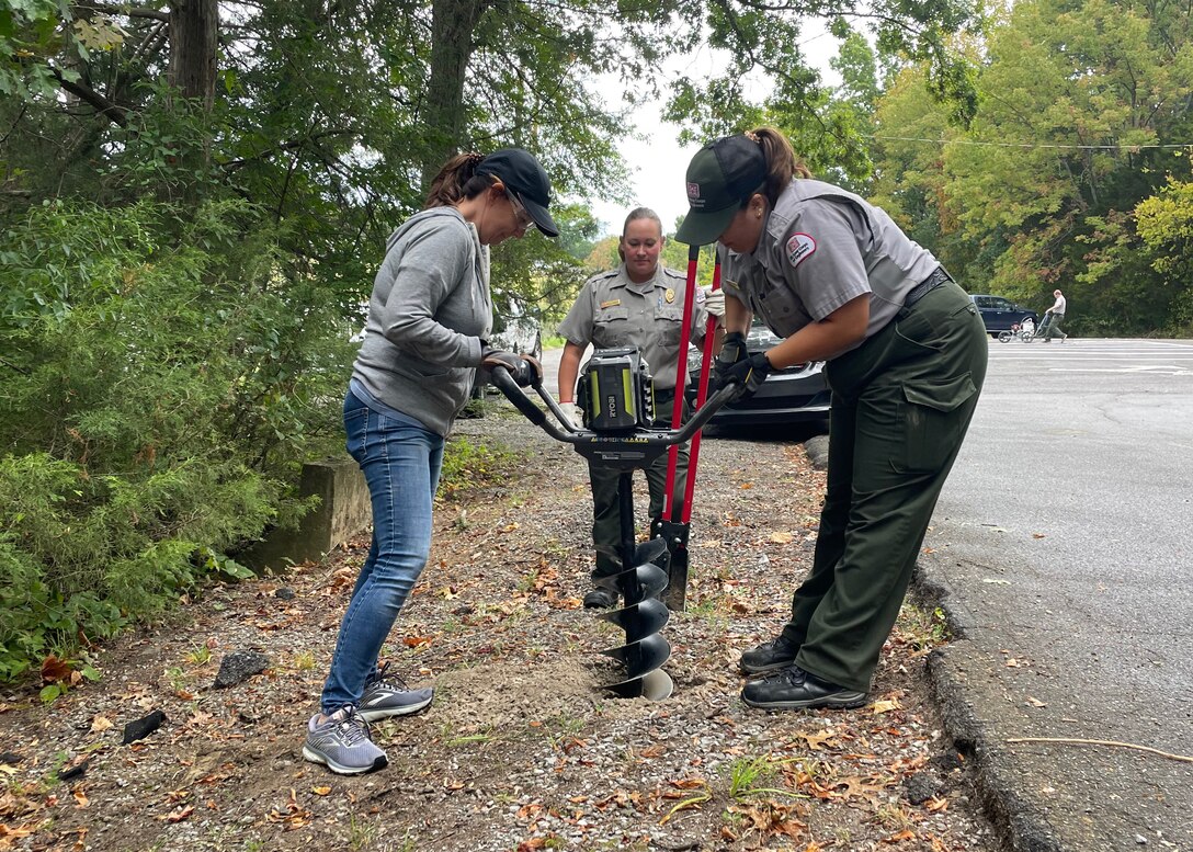 Park Ranger Danielle Knowles oversees Park Ranger Emily Johnson and volunteer Kim Johnson, as they use a large drill to create a new hole for a parking lot sign. The new signs periodically placed around the parking lot will help guide traffic and share important information with visitors at Stark Knob Boat Ramp on Old Hickory Lake in Hendersonville, Tennessee.