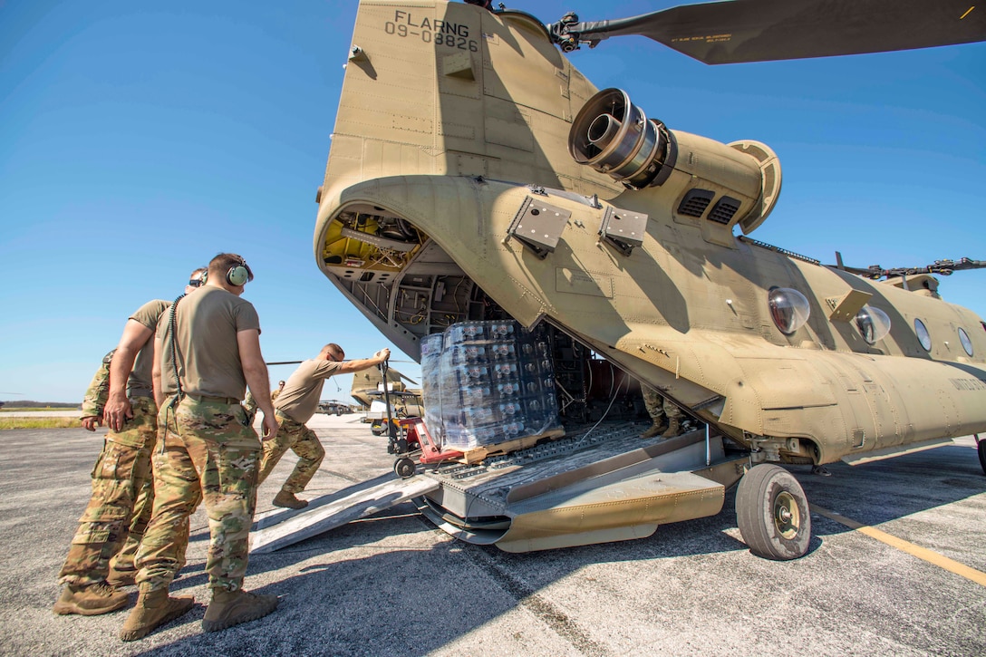 A National Guardsman load a pallet of water into an aircraft parked on a tarmac as two service members watch.