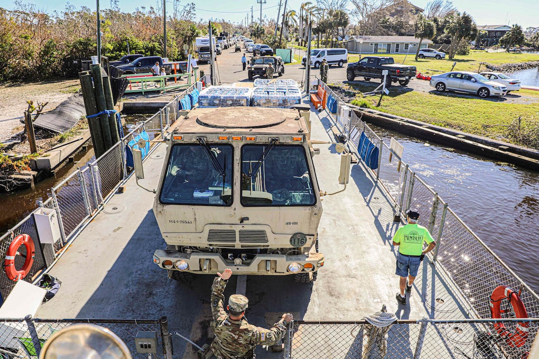 A service member directs a bus carrying supplies onto a boat.