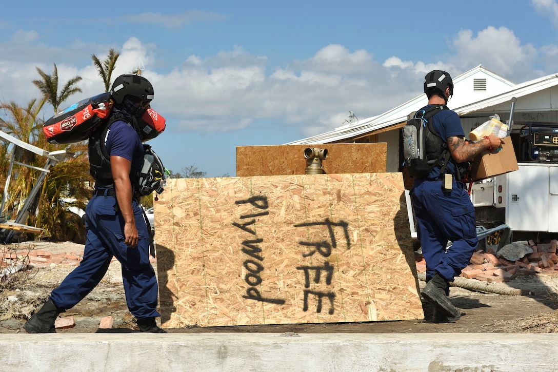 Coast Guardsmen carry boxes of supplies near debris.