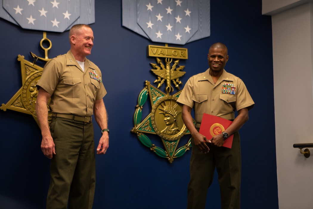 Two Marines standing in front of a blue curtain, smiling. The man on the right is holding an award.