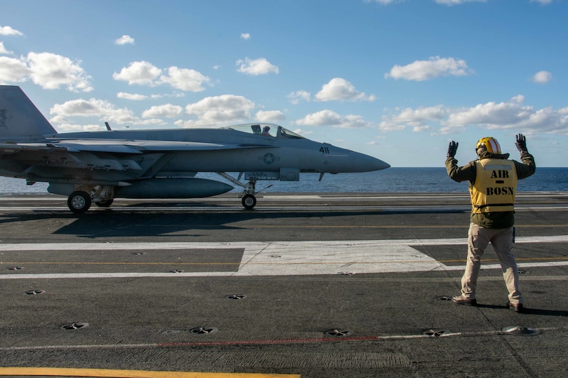 A service member wearing a yellow vest, signals to a pilot in a jet.