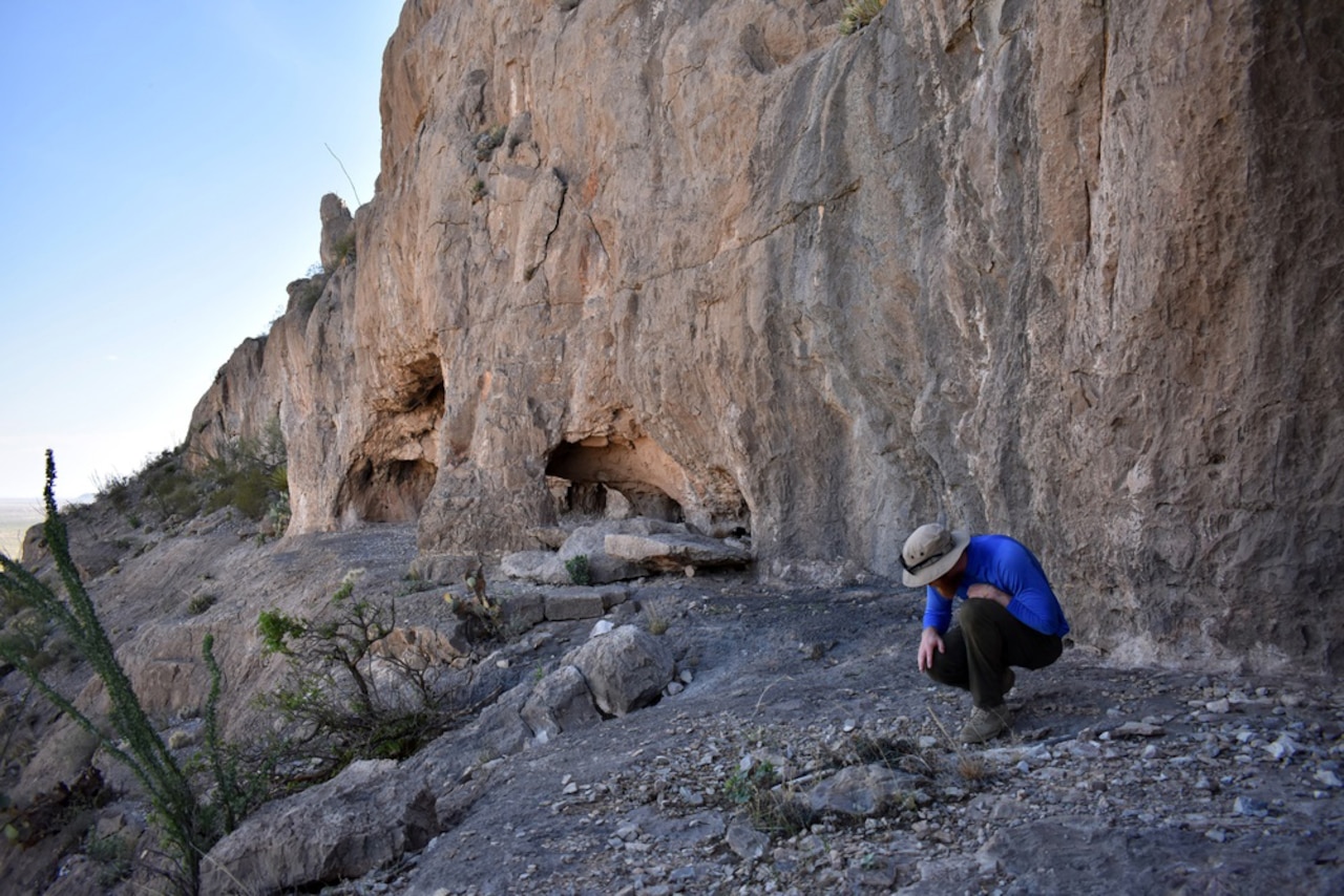 A man wearing a blue shirt looks at the ground at the base of a hill.