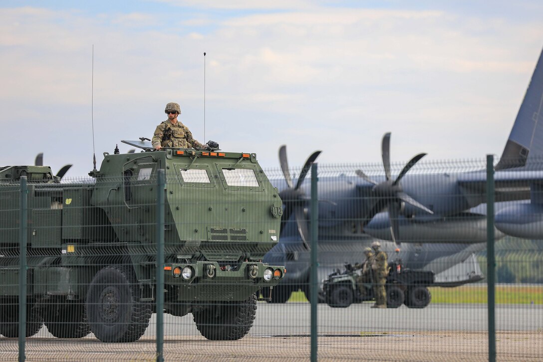 A soldier sits on top on a moving vehicle behind a wire fence.