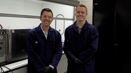 Two men wearing safety glass stand next to each other and smile into camera lens.