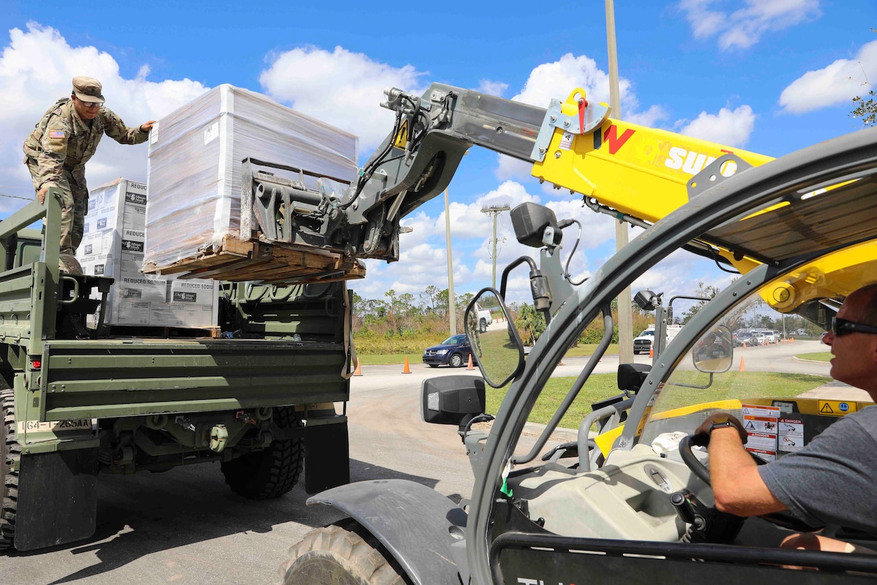 A soldier stands in the back of a truck as another person loads in cargo.