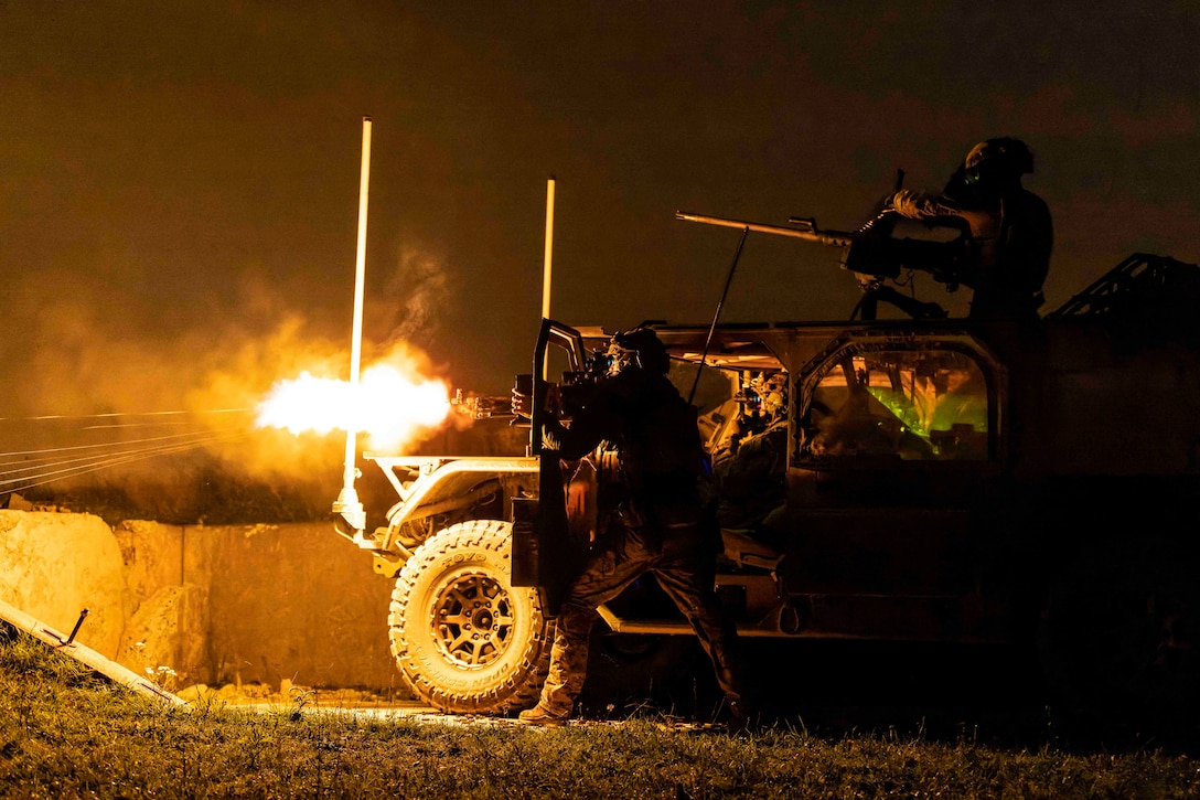 A soldier fires a weapon from behind a vehicle door as another soldier mans a gun on top of the vehicle at night.