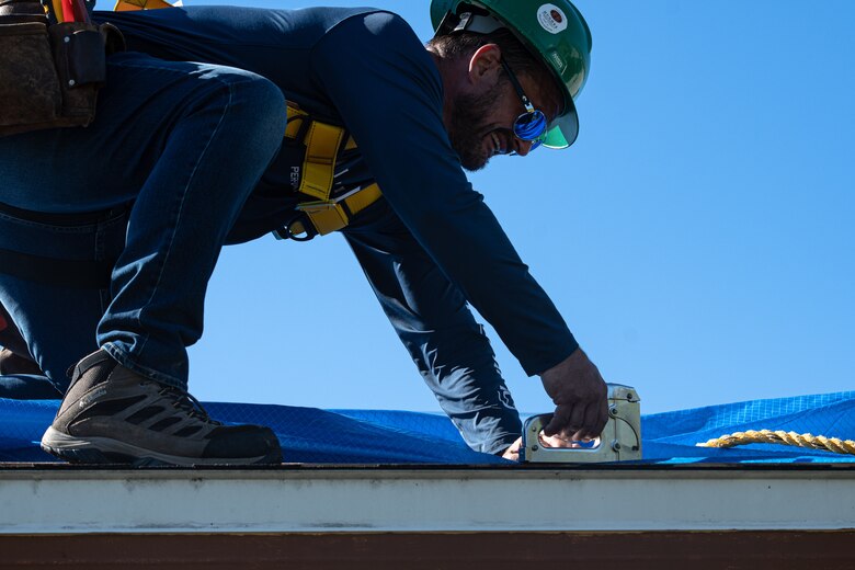 A worker uses staples to secure a blue tarp to a roof