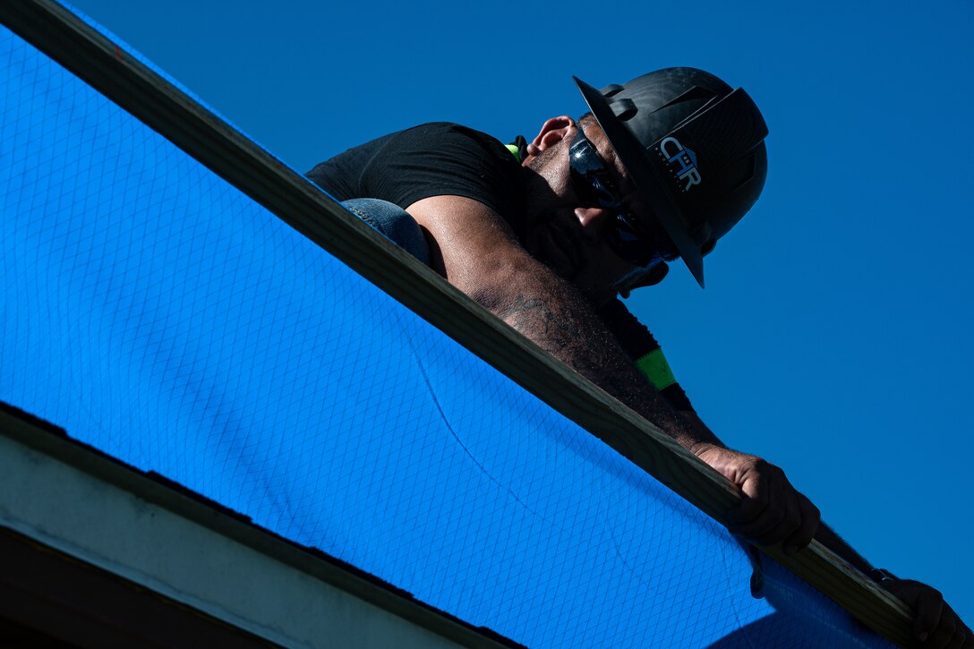 Worker stretches blue tarp across a roof.