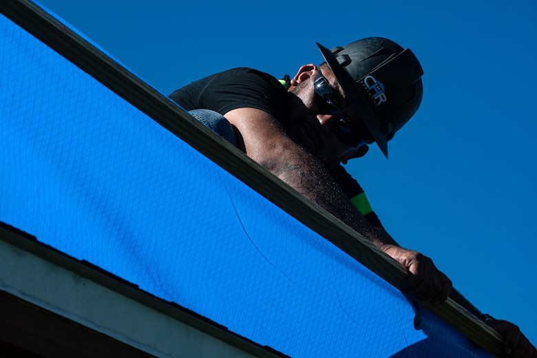 Worker stretches blue tarp across a roof.