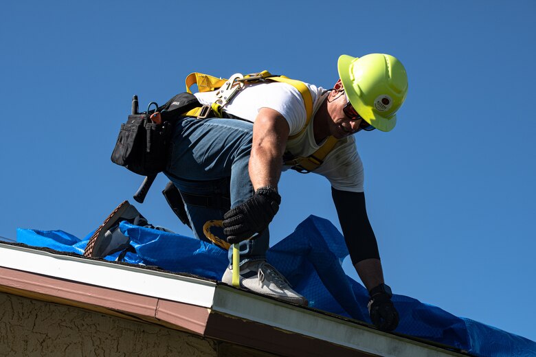 Worker in a hard hat secures blue tarp to a roof using hammer and nails
