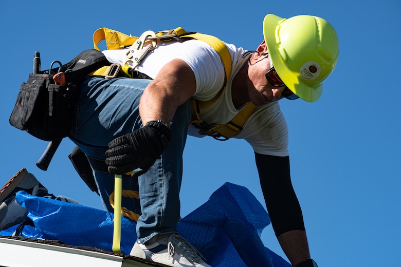 Roofer uses hammer and nails to secure a blue tarp to the shingles of a roof.