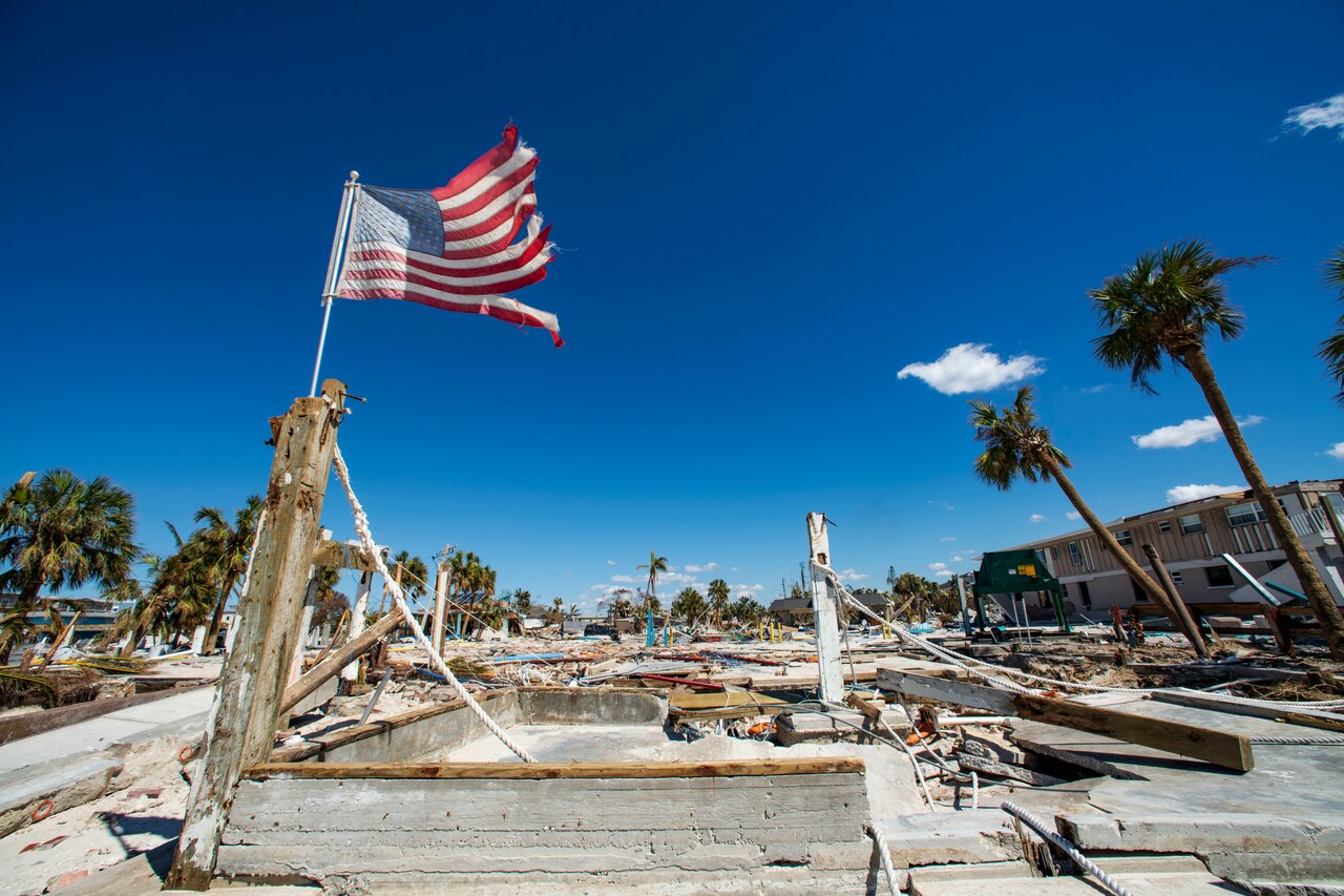 Rubble surrounds a tattered American flag as it waves in the air.