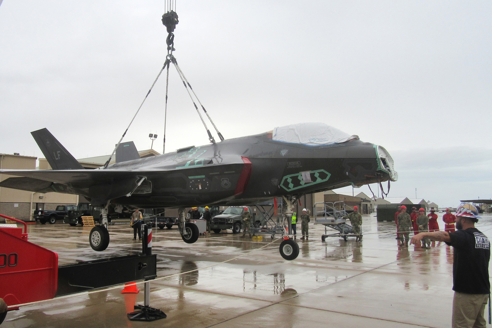 A reassembled F-35 is hoisted in the air by a crane cable while students observe.