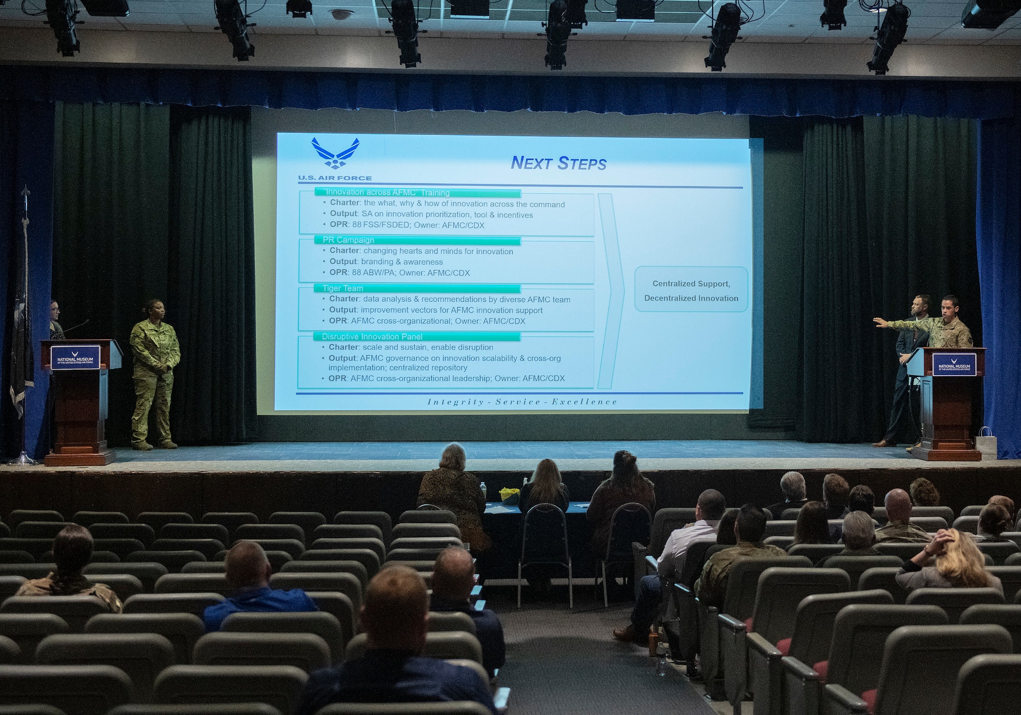 Two program graduates stand on stage presenting their capstone to classmates. Slide reads "Next Steps".