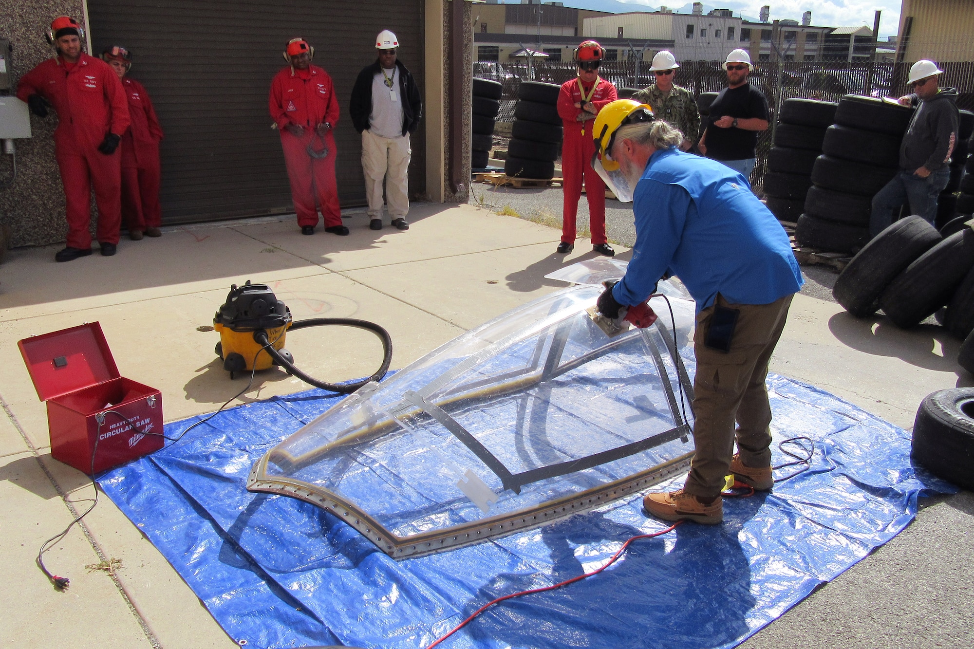 A student uses a cutting device on an aircraft canopy sitting on the ground while others observe.