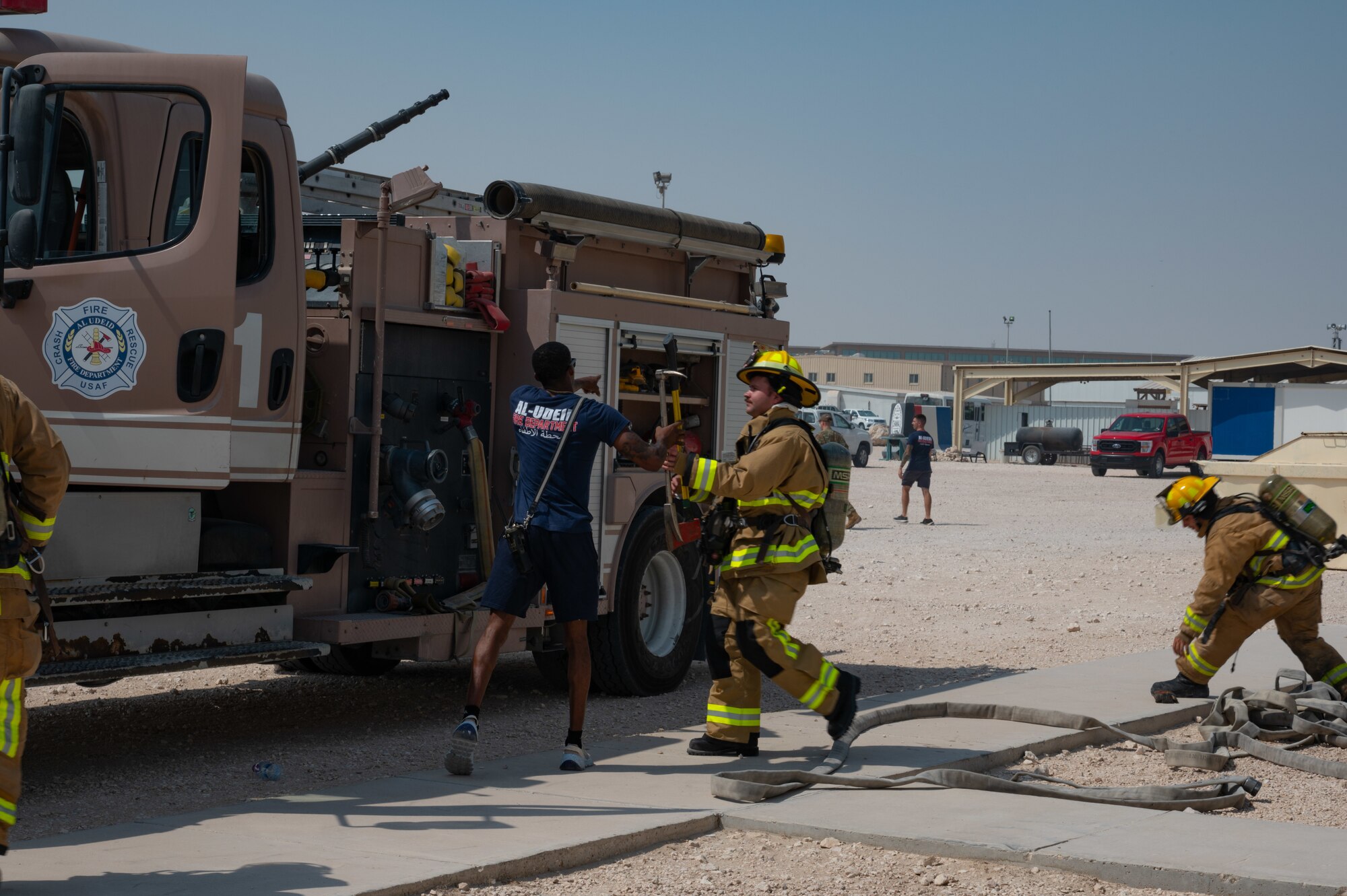 U.S. Air Force firefighters assigned to the 379th Expeditionary Civil Engineer Squadron (center) exchange a flat head axe and halligan bar while another firefighter assists in stretching out the firehose (right) during an exercise Aug. 30, 2022 at Al Udeid Air Base, Qatar. The tools exchanged between driver and operator were used to forcibly enter sealed rooms within the structure during the exercise. (U.S. Air Force photo by Staff Sgt. Dana Tourtellotte)