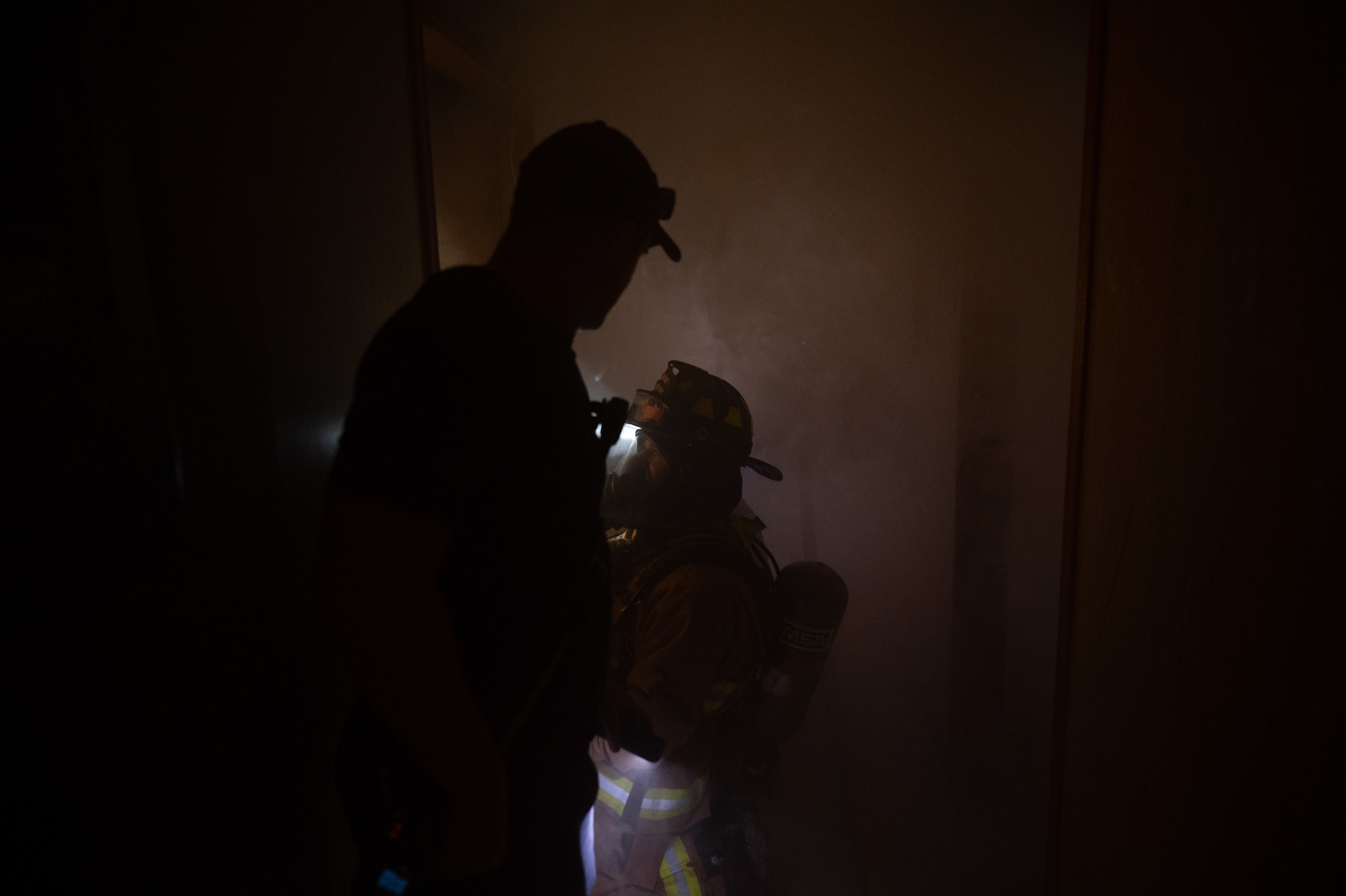 U.S. Air Force Staff Sgt. Logan Williams, part of the firefighter training team looks on as Senior Airman William Webber, a firefighter assigned to the 379th Expeditionary Civil Engineer Squadron, stays low to keep cool while opening a door during an exercise Aug.30, 2022 at Al Udeid Air Base, Qatar. The firefighters entered a simulated smoke filled facility to remain proficient in conducting search and rescue operations in low visibility environments. (U.S. Air Force photo by Staff Sgt. Dana Tourtellotte)